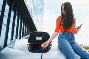 Using smartphone while waiting. Woman on the electric cars charge station at daytime. Brand new vehicle. photo