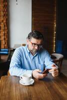 tomando tiempo para el descanso para tomar café. hombre maduro confiado en ropa formal tomando café y escribiendo un mensaje en el teléfono móvil mientras está sentado en el restaurante foto