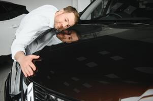 Happy handsome bearded man buying a car in dealership, guy hugging hood of new car photo
