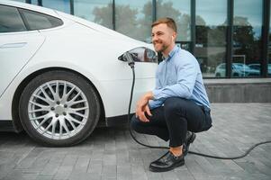 Hansome bearded guy sitting near his new modern electric car and holding plug of the charger, while car is charging at the charging station. photo