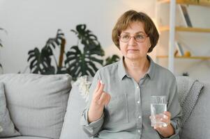 age, medicine, healthcare and people concept - senior woman with glass of water taking pills at home photo