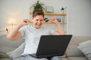 Happy excited grey haired mature woman celebrating online win, using laptop, looking at screen, sitting on couch at home, middle aged female feeling amazed, surprised by unbelievable good news. photo