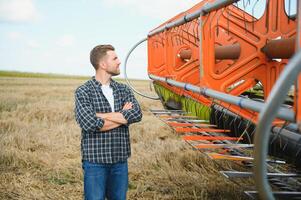 Happy farmer proudly standing in a field. Combine harvester driver going to crop rich wheat harvest. Agronomist wearing flannel shirt, looking at camera on a farmland photo