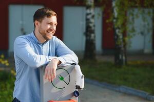 Portrait of a young man standing with charging cable near the charging station. Concept of fast home car chargers photo