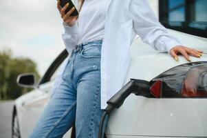 Phone in hands. Woman on the electric cars charge station at daytime. Brand new vehicle. photo