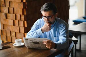 Closeup portrait of serious senior handsome man reading newspaper, having coffee break and sitting at table. photo