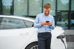 Handsome man in business suit surfing internet on modern smartphone while waiting electric car to charge. photo