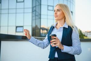 hermosa mujer yendo a trabajo con café caminando cerca oficina edificio. retrato de exitoso negocio mujer participación taza de caliente beber. foto