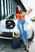 utilizando teléfono inteligente mientras espera. mujer en el eléctrico carros cargar estación a tiempo de día. marca nuevo vehículo. foto