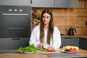 contento sonriente linda mujer es preparando un Fresco sano vegano ensalada con muchos vegetales en el cocina a hogar y molesto un nuevo receta foto