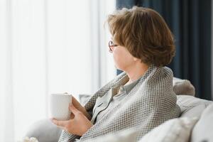 Smiling mature elder 65s woman sitting relaxing with cup of tea, coffee. Senior mid age stylish look woman with eyeglasses portrait with cup looking away at modern home photo
