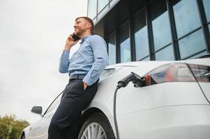 Handsome man in business suit surfing internet on modern smartphone while waiting electric car to charge. photo