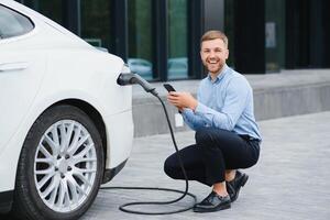 Hansome bearded guy sitting near his new modern electric car and holding plug of the charger, while car is charging at the charging station. photo