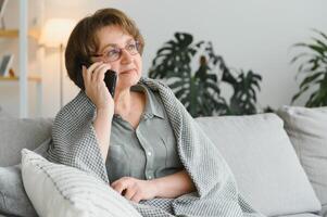 bella anciana con ropa informal usando su teléfono inteligente mientras se sienta en el sofá de casa foto