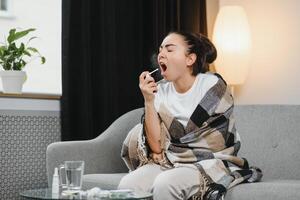 sick young woman in warm clothes spraying throat while sitting on couch at home. photo