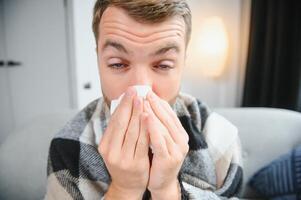 joven hombre sufrimiento desde un común frío y gripe o alergia sentar a hogar envuelto en cobija y limpiar su nariz con tejidos mientras él tener un fuerte dolor de cabeza dolor, cuidado de la salud concepto. foto