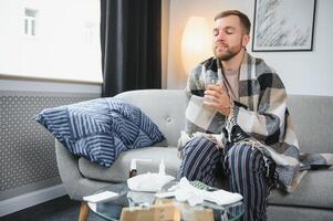 Sick bearded man who has bad cold or seasonal flu sitting on couch at home. Guy with fever wearing warm plaid shivering with worried face expression. photo