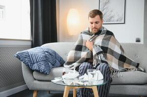 enfermo barbado hombre quien tiene malo frío o estacional gripe sentado en sofá a hogar. chico con fiebre vistiendo calentar tartán temblando con preocupado cara expresión. foto