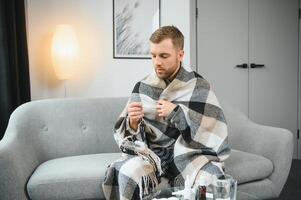 Sick bearded man who has bad cold or seasonal flu sitting on couch at home. Guy with fever wearing warm plaid shivering with worried face expression. photo