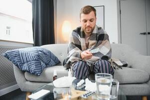 un enfermo hombre se sienta a hogar en un gris sofá con un frazada. enfermedad, proteccion, coronavirus, enfermedad, gripe foto