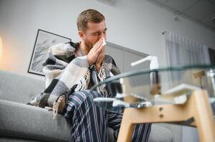 Young man suffering from a common cold and flu or allergy sit at home wrapped in blanket and wipe his nose with tissues while he have a strong headache pain, healthcare concept. photo