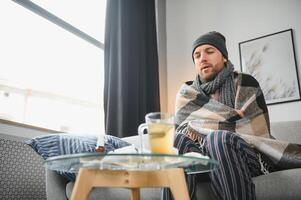 es frío a hogar en invierno hombre congelación en su casa en invierno porque de roto termostato. joven chico envuelto en de lana tartán temblando mientras sentado en sofá en vivo habitación interior foto