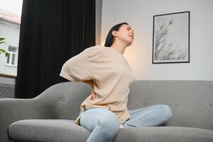 Upset mature woman suffering from backache, unhappy senior blonde sitting on a sofa at living room, feeling discomfort because of pain in back photo