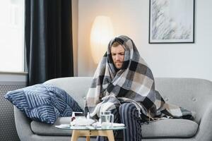 Sick adult man who has bad cold or seasonal flu virus is fighting illness at home. Unhappy middle aged man is sitting on sofa wrapped in warm scarf and plaid, holding paper tissue and sneezing. photo