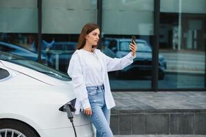 Happy young adult woman smiling wide, looking away, charging automobile battery from small public station, standing near electric car. photo