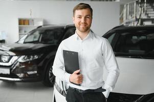 asistente en vehículo buscar. retrato de un hermoso joven coche ventas hombre en Ropa formal participación un portapapeles y mirando a cámara en un coche concesión foto