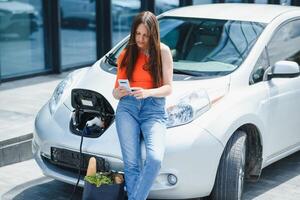 Using smartphone while waiting. Woman on the electric cars charge station at daytime. Brand new vehicle. photo