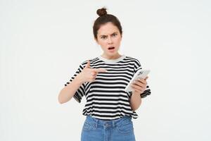 Image of angry young woman complains, points at telephone and looks disappointed, isolated against white background photo