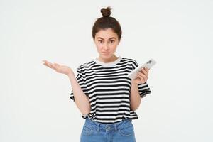 Portrait of shocked girl shrugs shoulders, looks confused reaction to something she saw on mobile phone, stands over white background photo