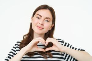 cerca arriba retrato de linda joven mujer muestra corazón firmar, mirando a cámara con amor y cuidado, en pie terminado blanco antecedentes foto