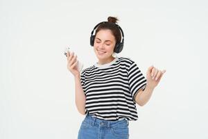 niña con teléfono inteligente baile, escuchando a música en inalámbrico auriculares, utilizando móvil teléfono transmisión aplicación a disfrutar favorito canciones, aislado terminado blanco antecedentes foto