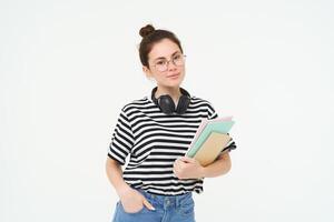 Education concept. Smiling brunette girl, student in casual clothes, holds her books, study material, wears headphones over neck, looks confident and happy, isolated over white background photo