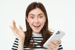 retrato de feliz, sorprendido joven mujer, participación móvil teléfono, demostración asombrado reacción a genial noticias, en pie con teléfono inteligente terminado blanco antecedentes foto