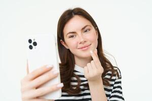 Portrait of beautiful woman showing heart sign and posing for selfie, taking picture of herself on smartphone app, posing near something cute, white background photo