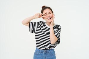 Cute european woman posing for photo, holding peace, kawaii hand gesture near face, smiling at camera, standing over white background photo