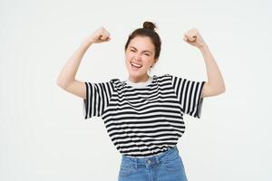 Woman power and feminism. Young girl feeling empowered and strong, flexing her biceps, showing muscles on arms, standing over white background photo