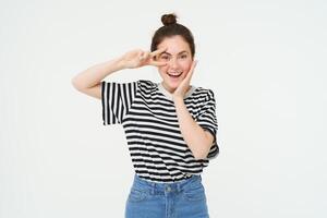 Beautiful brunette woman shows peace sign near eye and smiling, isolated against white background photo