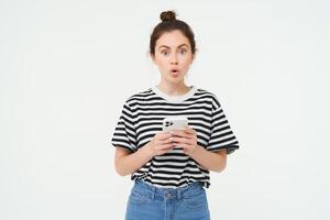 Portrait of impressed girl looks amazed, gasping, holding mobile phone, standing against white background photo