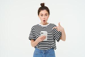 retrato de impresionado mujer, gotas mandíbula, puntos a su teléfono inteligente, ve increíble Noticias en línea en móvil teléfono, aislado terminado blanco antecedentes foto