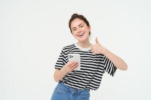 Enthusiastic girl with smartphone shows thumbs up, isolated over white background photo