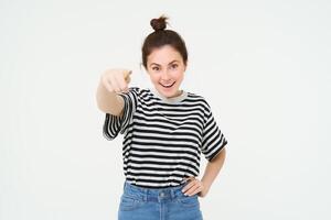 Image of confident, happy young woman in casual clothes, pointing finger at camera, laughing and smiling, standing over white background photo