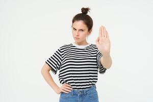 Image of beautiful woman with serious face, showing stop sign, extends one palm and frowns, stands over white background photo