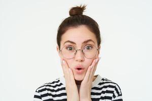 imagen de sorprendido linda niña en anteojos, sostiene manos en rostro, dice Guau, impresionado por algo, aislado terminado blanco antecedentes foto