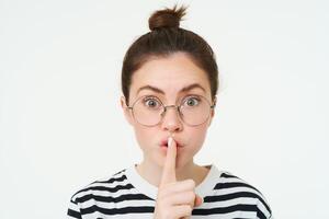 Portrait of girl with glasses, looks surprised, hughing, press finger to lips, shush shh gesture, asks to keep quiet, stands over white background photo