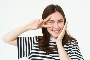 Portrait of beautiful, smiling female model, touches her clear glowing skin, shows peace sign and looks happy at camera, isolate on white background photo