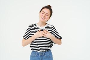 Image of beautiful woman in casual clothes, holds hands on heart and smiles, express gratitude, being thankful, stands over white background photo